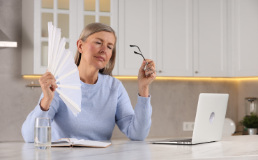 A middle age woman sitting at her desk fanning herself due to menopausal hot flushes